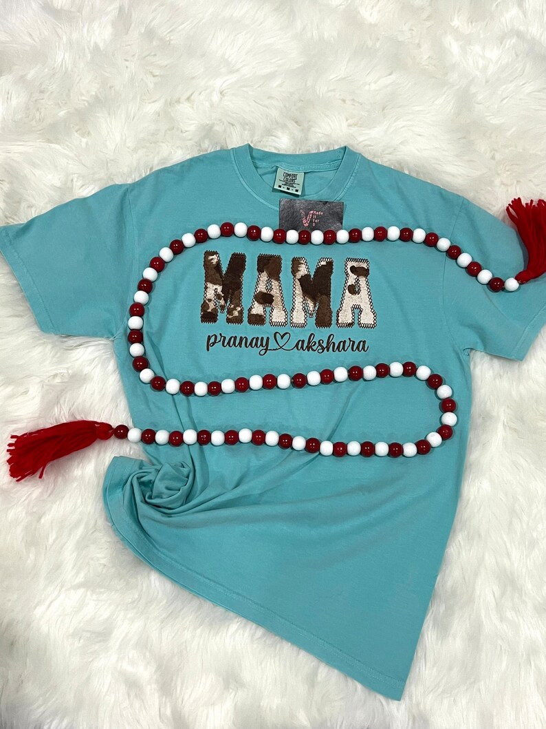 May include: A light blue t-shirt with a brown and white "MAMA" graphic with a cow print design. The shirt also has the text "pranay akshara" in brown lettering with a heart symbol between the words.