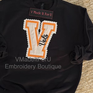 May include: Black sweatshirt with a large, orange and white letter "V" applique. The letter is outlined with a white lace trim and the word "Vols" is embroidered on the letter. The text "V Made It For U" is on a small tag.