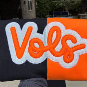 May include: A two-toned item, split vertically between black and orange. The word "Vols" is embroidered in orange with a white outline and a light blue cloud-like background. The item appears to be a piece of apparel.