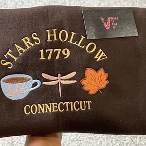 May include: Brown sweatshirt with embroidered text "Stars Hollow 1779 Connecticut" and images of a coffee cup, dragonfly, and maple leaf.