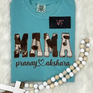 May include: A light blue t-shirt with a brown and white cow print design that says "MAMA" with a heart and the text "pranay akshara" underneath.
