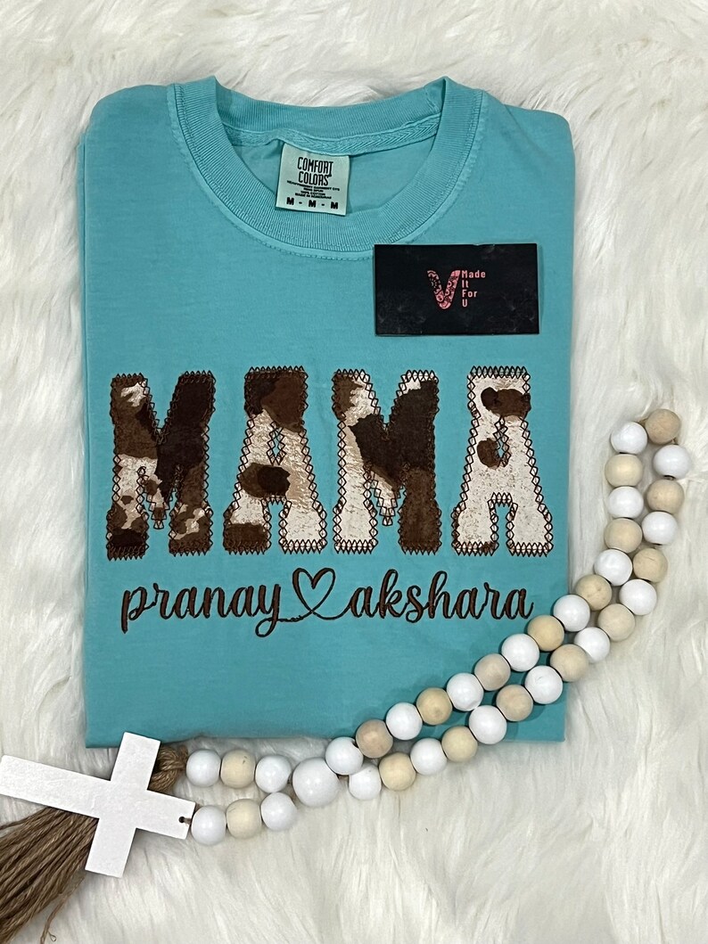May include: A light blue t-shirt with a brown and white cow print design that says "MAMA" with a heart and the text "pranay akshara" underneath.