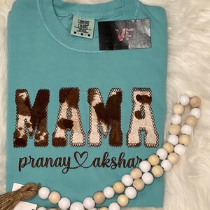 May include: A light blue t-shirt with the word "MAMA" in brown and white cow print lettering. The text "pranay akshar" is embroidered below the word "MAMA" in brown thread.