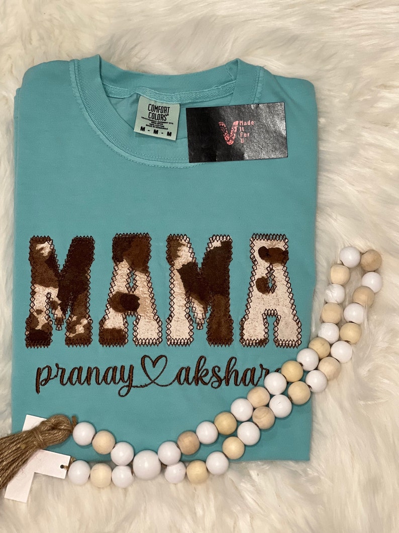 May include: A light blue t-shirt with the word "MAMA" in brown and white cow print lettering. The text "pranay akshar" is embroidered below the word "MAMA" in brown thread.