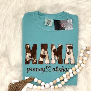 May include: A turquoise blue t-shirt with the word "MAMA" in brown and white cow print letters. The text "pranay akshar" is embroidered in brown below the word "MAMA".