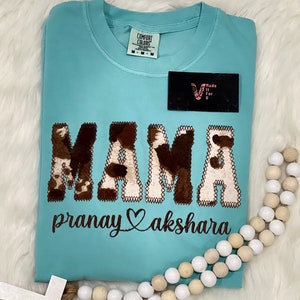 May include: A light blue t-shirt with a brown and white cow print "MAMA" design. The text "pranay akshara" is embroidered below the design.