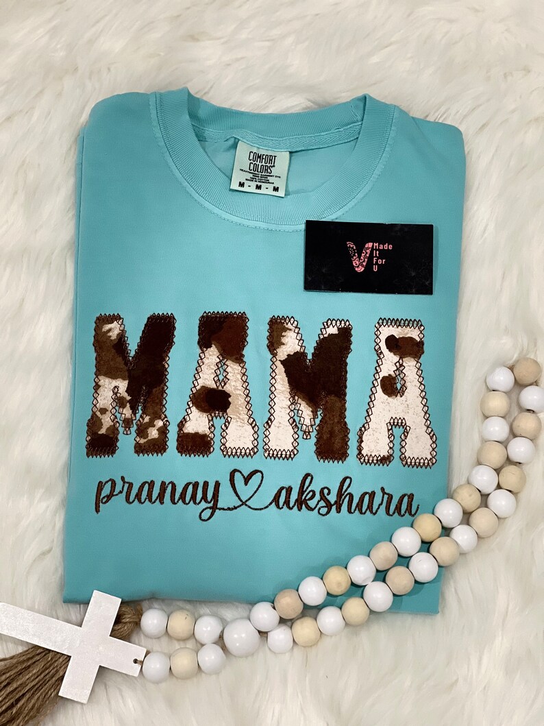 May include: A light blue t-shirt with a brown and white cow print "MAMA" design. The text "pranay akshara" is embroidered below the design.