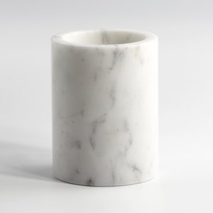 May include: A cylindrical marble container with a smooth, polished surface. The white stone features subtle gray veining throughout, creating a natural pattern. The top has a circular opening, and the base is flat. This decorative piece is suitable for various uses.