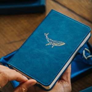 Puede incluir: Un diario encuadernado en cuero azul con un diseño de ballena dorada. El diario está cerrado y está siendo sostenido por una mano.