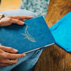 May include: A blue notebook with a gold whale design. The notebook is closed and being held by a person's hands. The notebook is sitting on a person's lap.