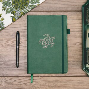 May include: A dark green leather-bound journal with a geometric turtle design in a light pink colour. The journal has a black elastic closure and a green ribbon bookmark.