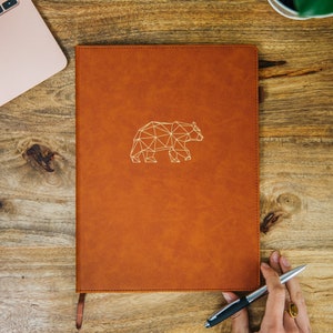 Może przedstawiać: Brązowy, skórzany notes z wytłoczonym geometrycznym wzorem niedźwiedzia na okładce. Notes leży na drewnianej powierzchni.