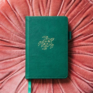Könnte beinhalten: Ein grünes Lederjournal mit einem goldenen geometrischen Schildkröten-Design auf dem vorderen Umschlag. Das Journal liegt auf einem rosa Samtkissen.