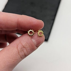 Peut inclure: Une paire de boucles d'oreilles en or en forme de soleil avec un petit cercle au centre. Les boucles d'oreilles sont petites et délicates.