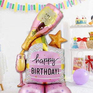 以下が含まれることがあります： シャンパンボトル、グラス、星、ギフトボックスが付いたピンクとゴールドのバルーンブーケ。ギフトボックスはピンクとグレーのヘリンボーン柄で、「ハッピーバースデー！」と書かれています。