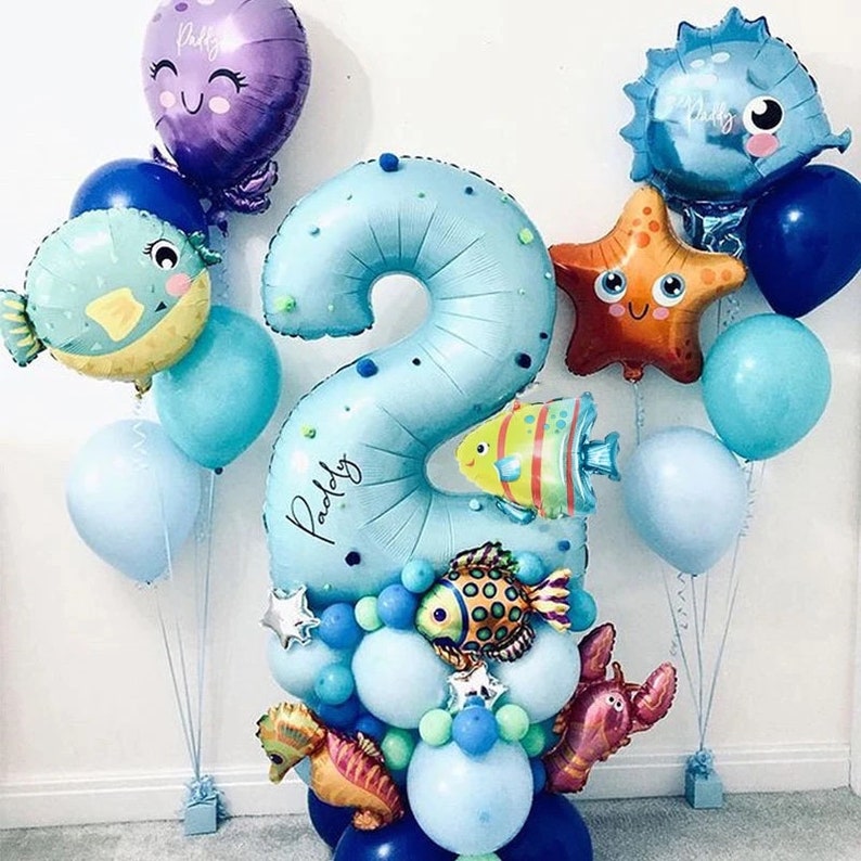 May include: A blue number two balloon with white polka dots and the name "Paddy" written on it. The balloon is surrounded by smaller blue, green, and orange balloons, as well as a starfish, a seahorse, and a pufferfish balloon.