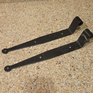 May include: Two black metal hinges with rounded ends and multiple holes. The hinges are designed for attaching a door or gate to a frame.