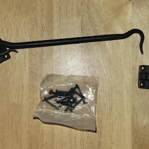 May include: Black metal door hook with a decorative scroll design. The hook is attached to a square plate with four screw holes. The hook is shown with a bag of screws.