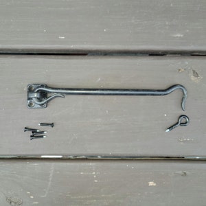Op de afbeelding: Zwarte metalen deurhaak met een vierkante basis en een gebogen haak. De haak is met schroeven aan een houten oppervlak bevestigd.