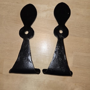 May include: Two black metal stirrups with a unique design. Each stirrup features a flared base, a central loop, and a teardrop-shaped top. The metal has a textured finish, suggesting a handcrafted appearance. These stirrups are likely for equestrian use.