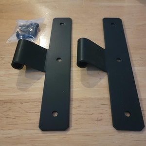 Puede incluir: Dos bisagras de metal negro con un diseño decorativo de desplazamiento. Las bisagras están unidas a una placa de metal plana.