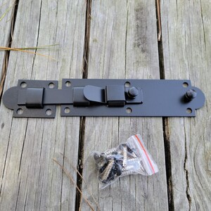 May include: A black metal gate latch with a sliding bolt mechanism, accompanied by a small bag of screws. The latch is rectangular with rounded edges and mounting holes, designed for secure closure. The hardware is set against a weathered wooden surface.