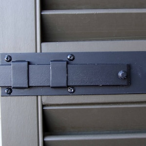 Può includere: Chiavistello per persiana in metallo nero con una barra rettangolare e un'estremità decorativa arrotondata. Il chiavistello è fissato a una persiana in legno con viti nere.