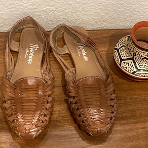 May include: A pair of brown leather huarache sandals with woven uppers and slingback straps, featuring the text Macarena Collection printed on the insoles, alongside a small ceramic pot with a black, white, and orange geometric pattern.