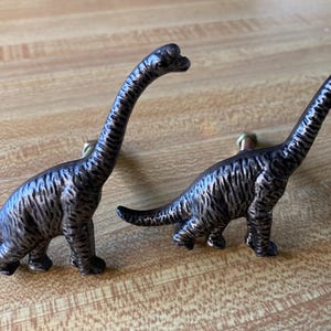 May include: Two dark bronze-colored metal cabinet knobs shaped like long-necked dinosaurs. Each knob features a textured surface and a small metal bolt protruding from the back for attachment to a drawer or cabinet door surface.