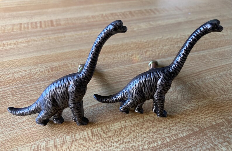 May include: Two dark bronze-colored metal cabinet knobs shaped like long-necked dinosaurs. Each knob features a textured surface and a small metal bolt protruding from the back for attachment to a drawer or cabinet door surface.