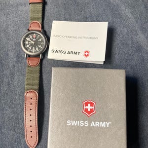 May include: A Swiss Army watch with a black dial, white numerals, and a green canvas strap with brown leather accents. The watch is displayed with its box and a booklet that reads "BASIC OPERATING INSTRUCTIONS". The box and booklet feature the Swiss Army logo.