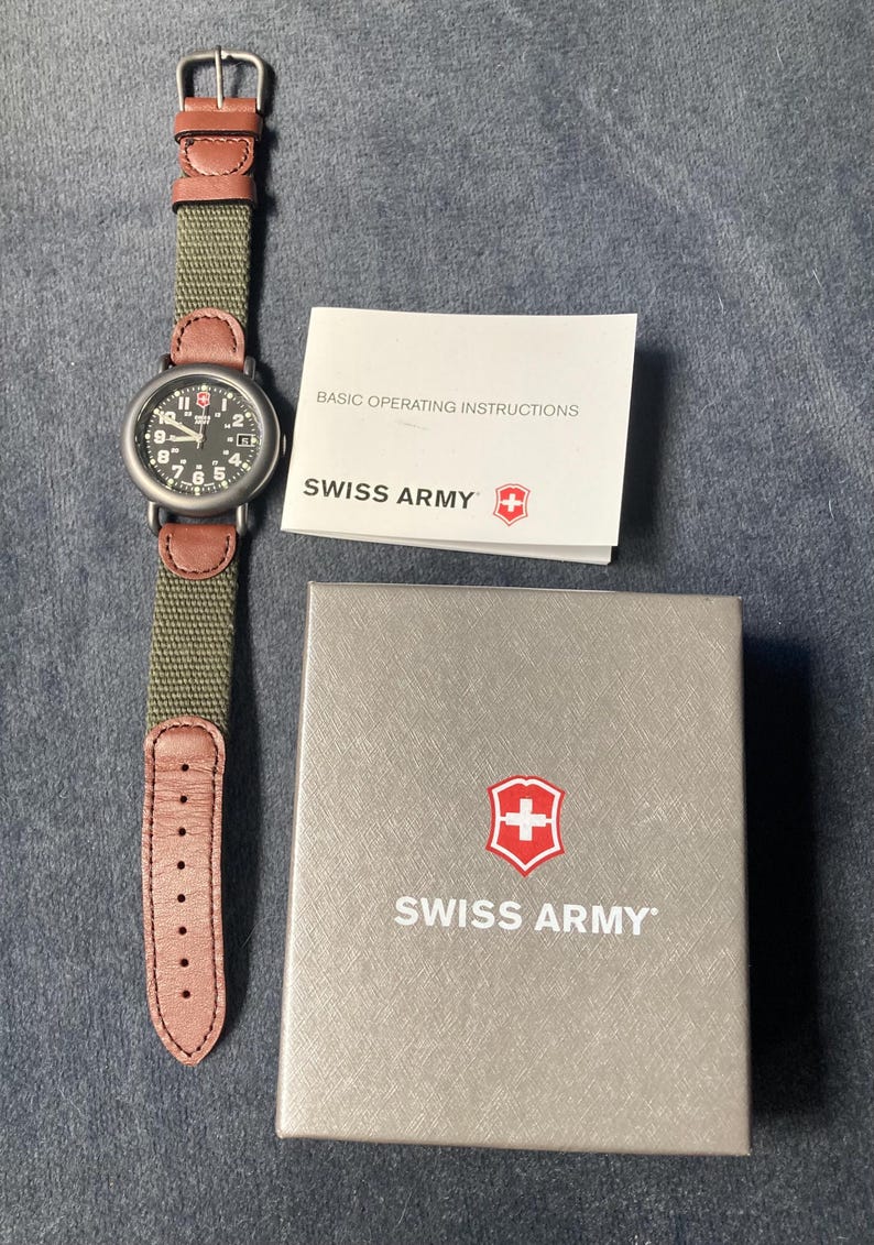 May include: A Swiss Army watch with a black dial, white numerals, and a green canvas strap with brown leather accents. The watch is displayed with its box and a booklet that reads "BASIC OPERATING INSTRUCTIONS". The box and booklet feature the Swiss Army logo.