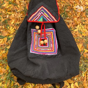 May include: A black fabric backpack featuring a flap with a multi-colored geometric pattern in red, blue, yellow, and green. The front pocket displays a square concentric design in matching colors, accented by red drawstrings and wooden beads.