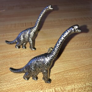 May include: Two silver-toned metal cabinet knobs shaped like long-necked dinosaurs. Each knob features a textured surface mimicking dinosaur skin, a long neck, a sturdy body, and a short tail, designed to be mounted onto furniture drawers.