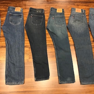 May include: Seven pairs of blue denim jeans laid out on a wooden floor. The jeans are all different shades of blue and have different levels of distressing.