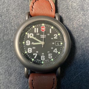 May include: A Swiss Army watch with a black face, white numerals, and a date window at the 3 o'clock position. The watch has a green canvas strap with brown leather accents. The watch face displays the Swiss Army logo.