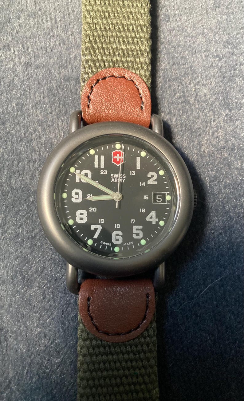 May include: A Swiss Army watch with a black face, white numerals, and a date window at the 3 o'clock position. The watch has a green canvas strap with brown leather accents. The watch face displays the Swiss Army logo.