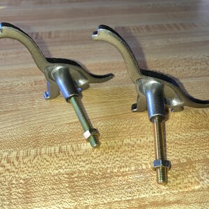 May include: Two metallic dinosaur-shaped drawer knobs with a textured gold-tone finish. Each knob features a long neck and tail, with a threaded metal bolt extending from the center of the body, secured by a small hexagonal nut at the end.