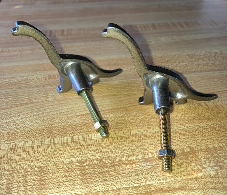 May include: Two metallic dinosaur-shaped drawer knobs with a textured gold-tone finish. Each knob features a long neck and tail, with a threaded metal bolt extending from the center of the body, secured by a small hexagonal nut at the end.