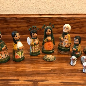 May include: A set of hand-painted wooden nativity figurines featuring various characters including figures in green, yellow, and orange robes, an angel, a central infant in a manger, and several small animal figures with floral details.
