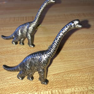 May include: Two metallic silver-toned dinosaur figurines shaped like long-necked sauropods. Each dinosaur features a textured surface with visible ridges along the body, a long neck, a small head, and a thick tail, standing in a side profile.