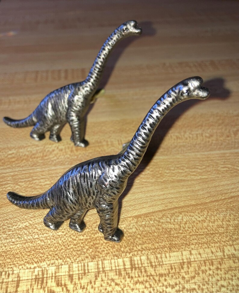 May include: Two metallic silver-toned dinosaur figurines shaped like long-necked sauropods. Each dinosaur features a textured surface with visible ridges along the body, a long neck, a small head, and a thick tail, standing in a side profile.