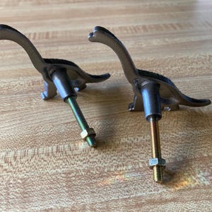May include: Two dark gray metal dinosaur-shaped cabinet knobs. Each knob features a long-necked sauropod silhouette with a threaded metal bolt extending from the back, complete with a small hexagonal nut attached to the screw for installation.