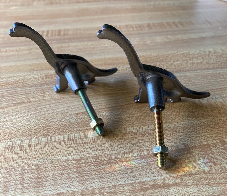 May include: Two dark gray metal dinosaur-shaped cabinet knobs. Each knob features a long-necked sauropod silhouette with a threaded metal bolt extending from the back, complete with a small hexagonal nut attached to the screw for installation.