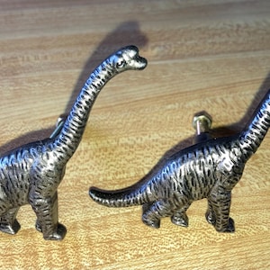 May include: Two metallic, silver-toned dinosaur-shaped cabinet knobs. Each knob features a long-necked sauropod design with a textured, brushed finish and a central mounting screw post extending from the body for attachment to furniture.
