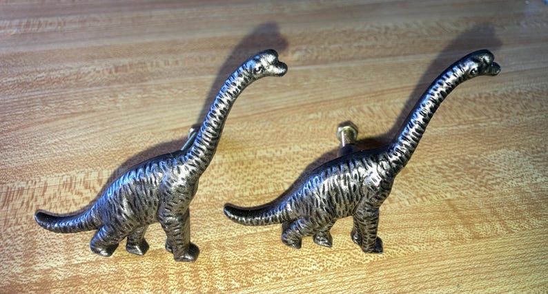 May include: Two metallic, silver-toned dinosaur-shaped cabinet knobs. Each knob features a long-necked sauropod design with a textured, brushed finish and a central mounting screw post extending from the body for attachment to furniture.