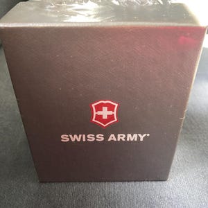 May include: A brown rectangular box with the words "SWISS ARMY" in white text. The Swiss Army logo, a white cross inside a red shield, is above the text. The box is wrapped in clear plastic.