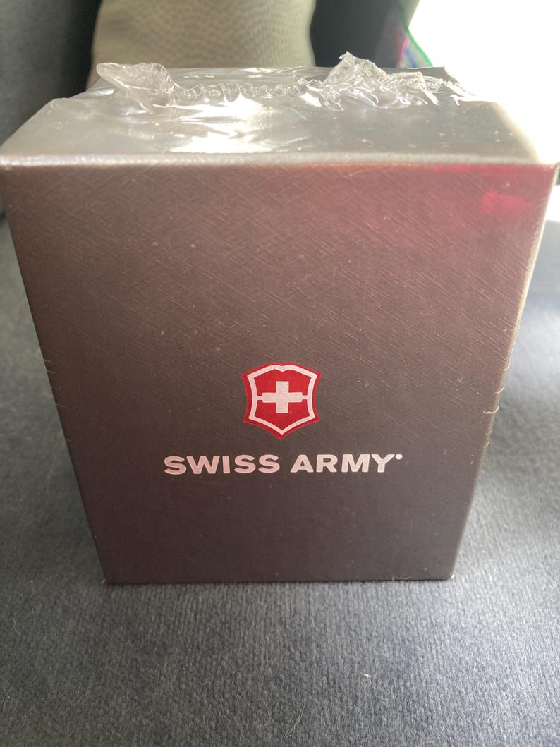 May include: A brown rectangular box with the words "SWISS ARMY" in white text. The Swiss Army logo, a white cross inside a red shield, is above the text. The box is wrapped in clear plastic.