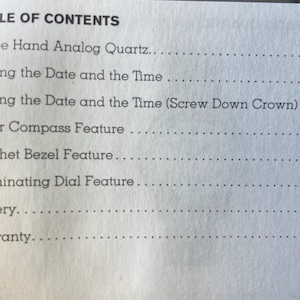 May include: A close-up of a printed page titled "TABLE OF CONTENTS." The text lists features of a watch, including "Three Hand Analog Quartz," "Setting the Date and the Time," and "Solar Compass Feature." The page is white with black text.