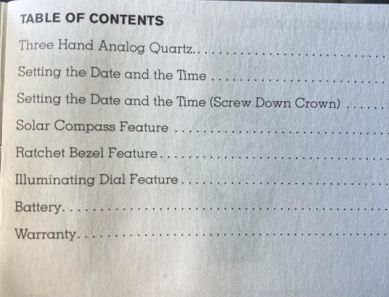 May include: A close-up of a printed page titled "TABLE OF CONTENTS." The text lists features of a watch, including "Three Hand Analog Quartz," "Setting the Date and the Time," and "Solar Compass Feature." The page is white with black text.